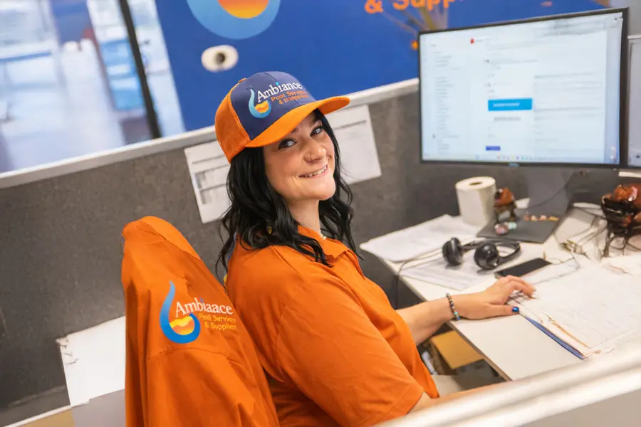 A person wearing an orange Ambiaace-branded shirt and hat sits at a desk with a computer monitor, keyboard, and office supplies, smiling at the camera in a work environment that fosters open communication.