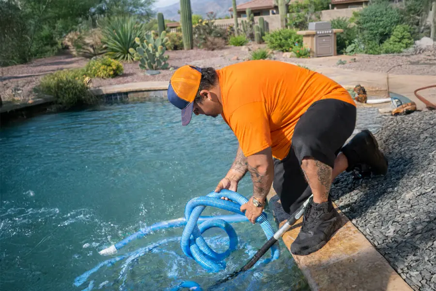 A person in an orange shirt and hat kneels by a backyard pool, engaged in pool maintenance by placing a blue hose in the water, surrounded by desert landscaping with cacti and rocks.