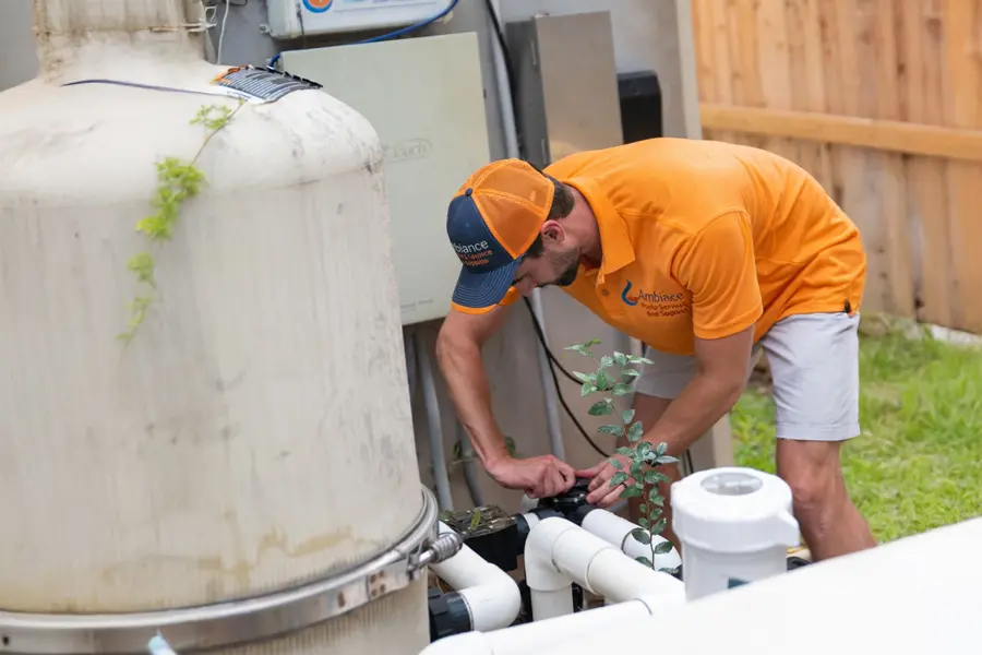 A man in an orange shirt and cap is inspecting or repairing white pipes connected to large equipment outdoors, demonstrating equipment care for pool protection near a wooden fence and grassy area.
