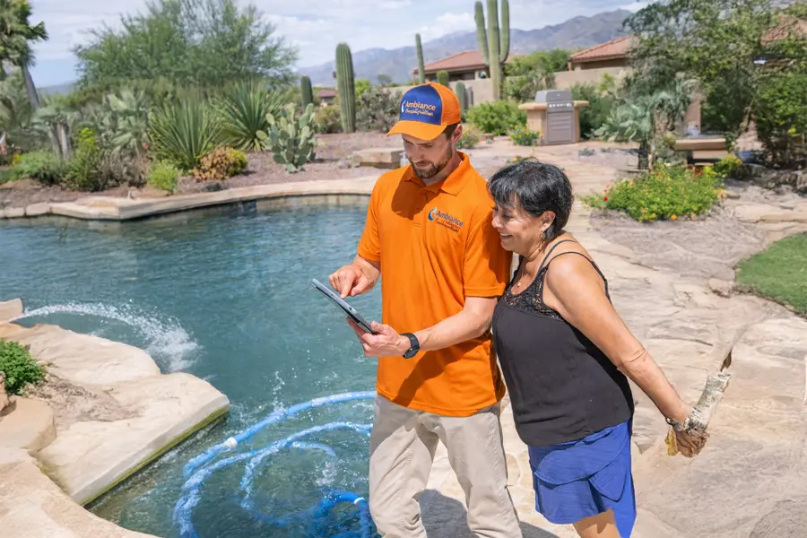A trustworthy technician in an orange uniform shows a tablet to a woman by an outdoor pool surrounded by desert landscaping, cacti, and mountains in the background on a sunny day.